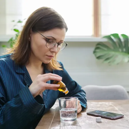 Nervous disorder, depression, nervousness, age-related health problems. Mature woman dripping drops with glass of water, drinking sedative medicine, at home in pajamas, copy space
